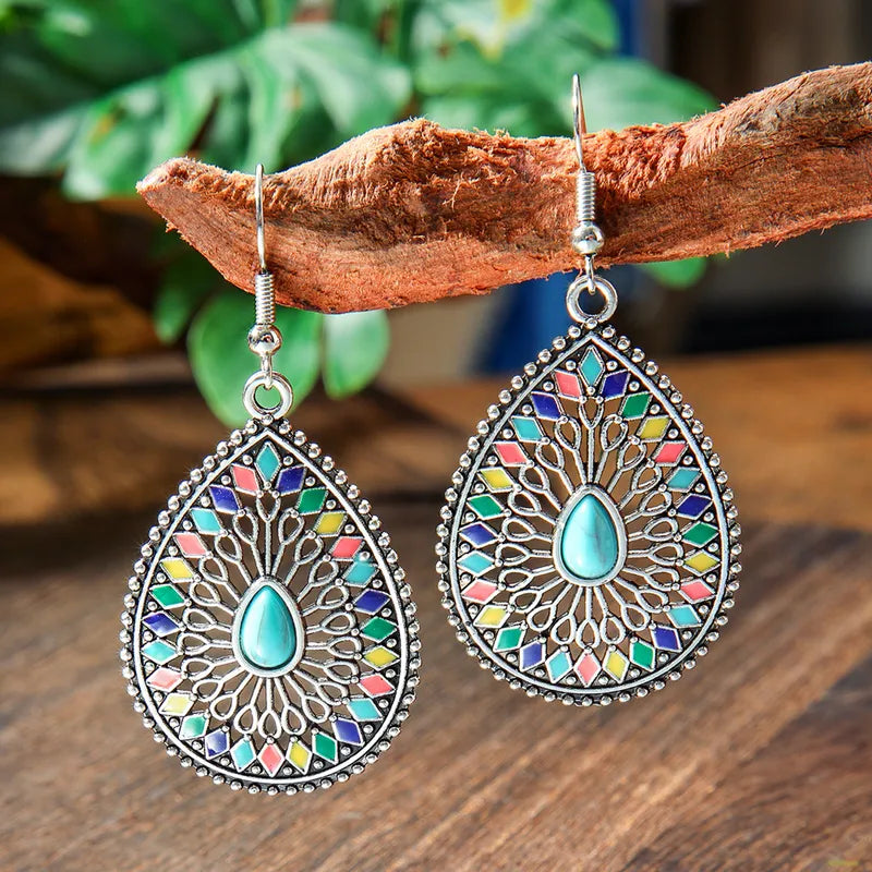 A pair of silver-tone teardrop drop earrings with central turquoise stones and colorful enamel hanging from a wooden branch.