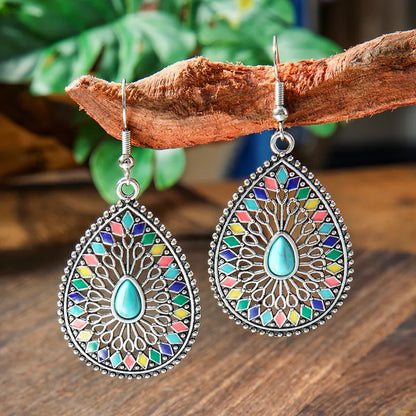 A pair of silver-tone teardrop drop earrings with central turquoise stones and colorful enamel hanging from a wooden branch.