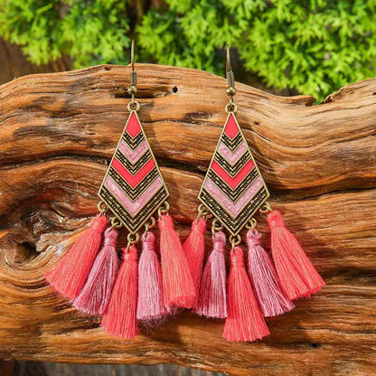 Pair of pink tassel earrings on a wooden surface with greenery in the background