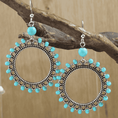 Front view of two large oxidised silver drop earrings with bright turquoise beads, suspended from a dark wooden branch agains