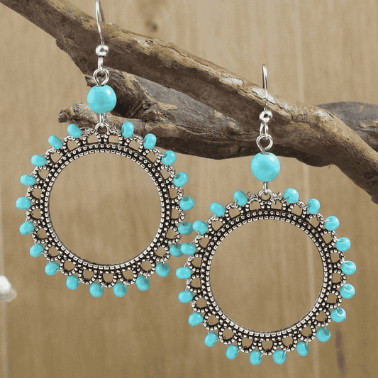 Front view of two large oxidised silver drop earrings with bright turquoise beads, suspended from a dark wooden branch agains