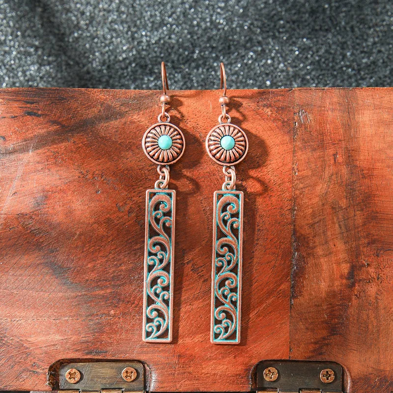 Pair of oxidised copper dangle earrings with round turquoise stones and rectangular filigree panels, resting on a rustic wood